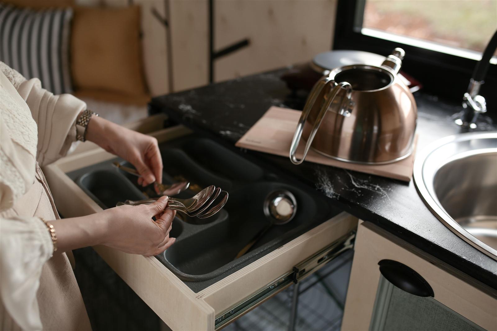 Person arranging spoons and utensils in an open kitchen drawer beside a stainless kettle