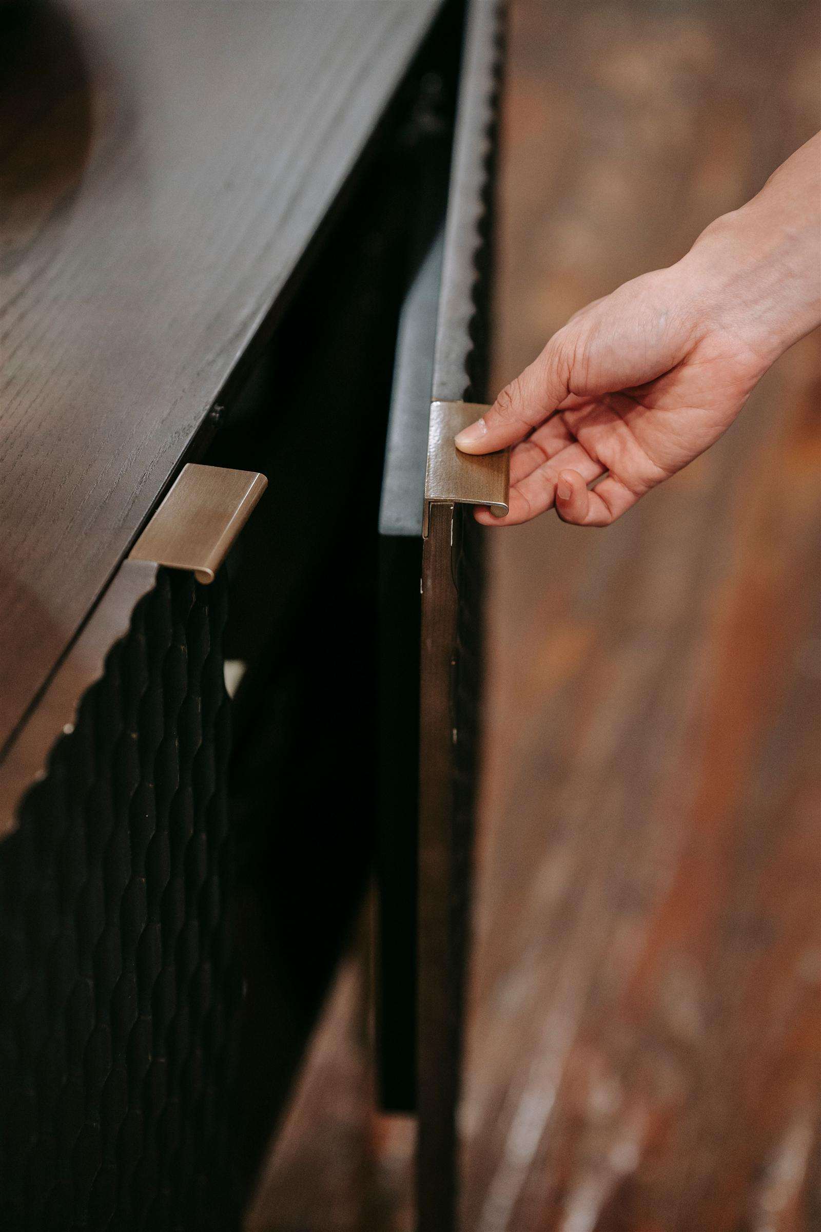 A hand gently gripping a brass handle to open a wooden cabinet door at home